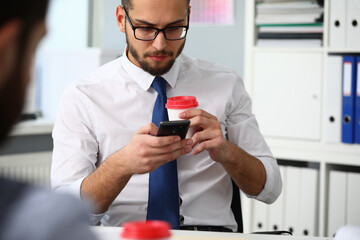 Young manager is spending break in work with hot drink and messaging on smart phone near colleague in office