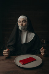 A beautiful nun is sitting at a table in front of a smartphone, holding a knife and a fork.