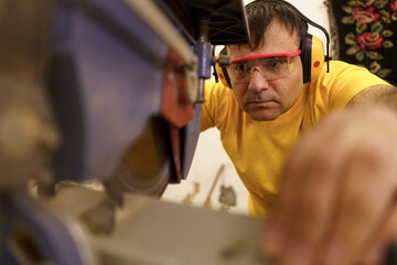 Carpenter cutting a piece of wood for furniture in his woodwork workshop, using a circular saw, and wearing safety goggles and earmuffs.