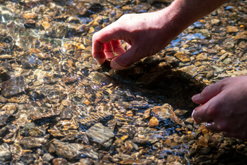 Male european hand refreshing in meditative zen atmosphere in a crystal clear water of a flat little creek with silky ripples and floating waves and sunny reflections searching for stones in the water