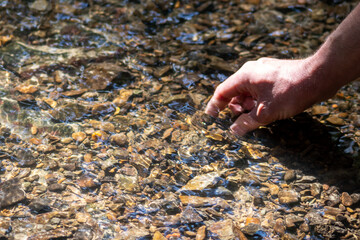 Male european hand refreshing in meditative zen atmosphere in a crystal clear water of a flat little creek with silky ripples and floating waves and sunny reflections searching for stones in the water