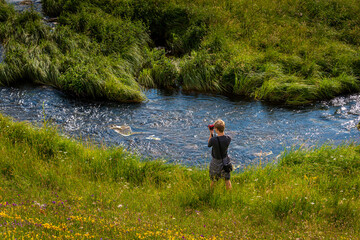 young man taking photos next to a river in the countryside, in summer .with wild flowers in forground.