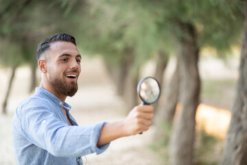 A young italian man bearded standing in a park with light jeans and a light blue shirt with a magnifying glass searches for something