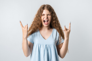 Emotive European young woman with curly long hair makes ROCK-N-ROLL sign, says: I will rock this party! yells loudly, wears casual t-shirt, stands over white background, feels self confident