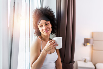 Woman in the morning. Attractive woman with sleeping mask is holding a cup with hot tea or coffee and looking at the sunrise standing near the window in her home and having a perfect cozy morning.