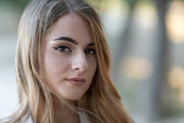 Fototapeta premium Charming Italian woman with long straight blonde hair and tight lips looks into the room in a park with trees