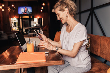 Smiling senior female blogger in casual wear sitting in cafe using laptop and smartphone.