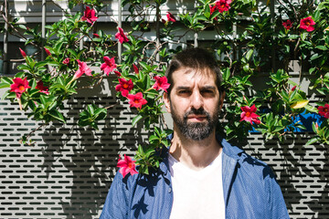 Bearded man in casual attire leaning on a flowered wall while looking camera