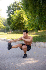 Obraz premium Full length portrait of a sportsman doing workout outside with naked torso, athletic body, sitting in knee, isolated on nature background. Healthy lifestyle concept. Vertical view.