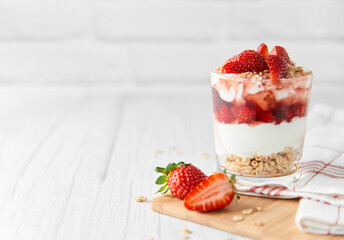 Homemade layered dessert with fresh strawberries, cream cheese or yogurt, granola and strawberry jam in glass on white wood background. Healthy organic breakfast or snack concept. Selective focus.