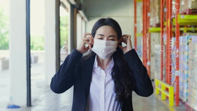 Asian young business woman in suit wearing protective face mask during walking in warehouse factory due to covid pandemic crisis. Girl working in logistic and storage industrial with care of health.