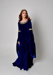 Naklejka premium Full length portrait of girl wearing long blue velvet gown with golden crown. standing pose with back to the camera, isolated against a studio background.