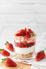 Homemade layered dessert with fresh strawberries, cream cheese or yogurt, granola and strawberry jam in glass on white wood background. Healthy organic breakfast or snack concept. Selective focus.