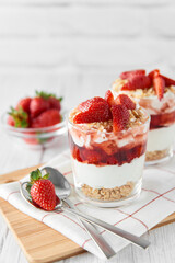 Homemade layered dessert with fresh strawberries, cream cheese or yogurt, granola and strawberry jam in glasses on white wood background. Healthy organic breakfast or snack concept. Selective focus.