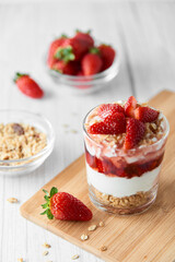 Homemade layered dessert with fresh strawberries, cream cheese or yogurt, granola and strawberry jam in glass on white wood background. Healthy organic breakfast or snack concept. Selective focus.