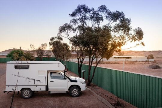 Small sized 4wd campervan parked on a camping at the desert. Sunset time. Green fence and tree next to the car. White vehicle. No logos. Coober Peddy, South Australia