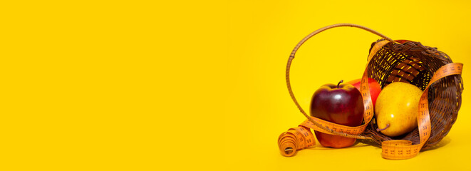 A basket of fresh fruit: a red Apple, a yellow pear and a mango lie on a yellow background, entangled with a measuring tape for the body: a healthy food concept, a banner with space for text