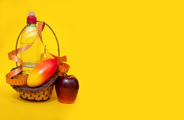 A basket of fresh fruit: a red Apple, a yellow pear and a mango lie surrounded by a measuring tape on a yellow background next to a sports water bottle: a healthy food concept, front view