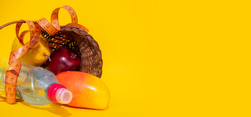 A basket of fresh fruit: a red Apple, a yellow pear and a mango lie surrounded by a measuring tape on a yellow background next to a sports water bottle: a healthy food concept,  place for text