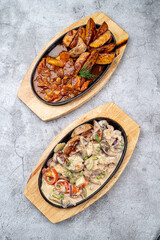 Top view of restaurant oven baked meals: vegetable ragout and meat with potato in spicy gravy, two cast-iron trays on grey concrete table