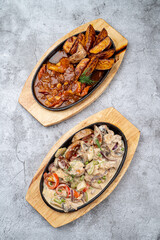 Top view of restaurant oven baked meals: vegetable ragout and meat with potato in spicy gravy, two cast-iron trays on grey concrete table