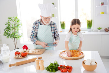 Photo of little girl granddaughter spend time with aged grandpa putting tomato sauce fresh cut ingredients dough family pizza recipe cooking together weekend home kitchen indoors