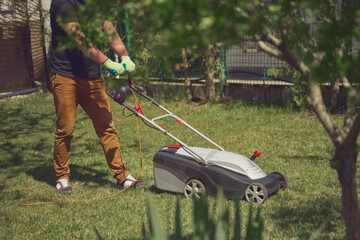 Young worker in casual clothes and gloves is mowing green grass with lawn mower on a yard. Gardening care equipment and services. Sunny day