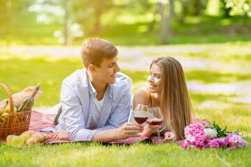 Fototapeta premium Romanic girlfriend with her boyfriend lying on picnic blanket and drinking wine at park on summer day