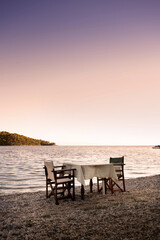 A table and two chairs stand on the beach near the Mediterranean sea in warm evening sun, waiting for a romantic dinner.