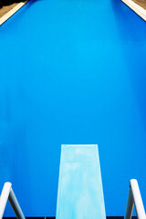 View from above to a light blue diving board and a blue pool with calm water surface in the sun.