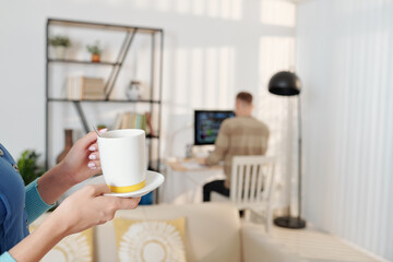 Fototapeta na wymiar Wife bringing cup of tasty coffee to husband who is working on programming code in background