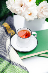 Cup of tea on books, flowers white wild rose in vase, warm plaid on white table outside in garden. Romantic leisure breakfast with nature background. Cozy home backyard or provence cafe