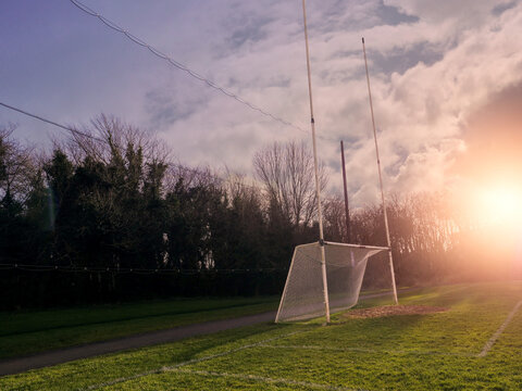 Green Fresh Grass In Focus. Irish National Sports Goal Posts Out Of Focus.Concept Practice Rugby, Hurling, Camogie, Gaelic Footbal. Sunset Time, Sun Flare. Selective Focus.