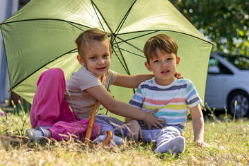 Portrait of happy small boy and girl with green umbrella.Brother and sister sitting in the garden and holding colorful umbrella. Time to relax,two kids having fun in the backyard, beautiful family day