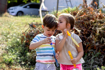 Three years old brother and sister blowing bubbles in the backyard.Cute blond girl kissing her little brother in the cheek.Children in the park blowing soap bubbles and having fun in the summer time.