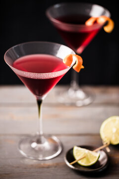 Closeup 2 glasses of cosmopolitan cocktail decorated with orange served with lime wedges.  Dark grey worktop with dark background