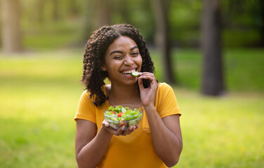 Pretty black girl eating fresh vegetable salad and winking at green park on summer day