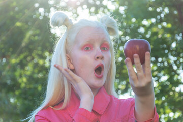 An albino girl in a red blouse against a background of green leaves and sunlight. she holds a red Apple in her hands and looks at the camera in surprise.