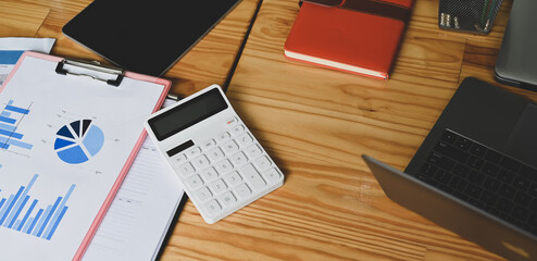 Top of a wooden working desk is surrounding by a computer laptop, paperwork, and office equipment.