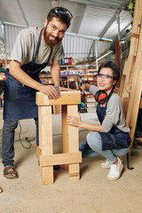 Cheerful young carpenters polishing surface of wooden stand they made with emery paper