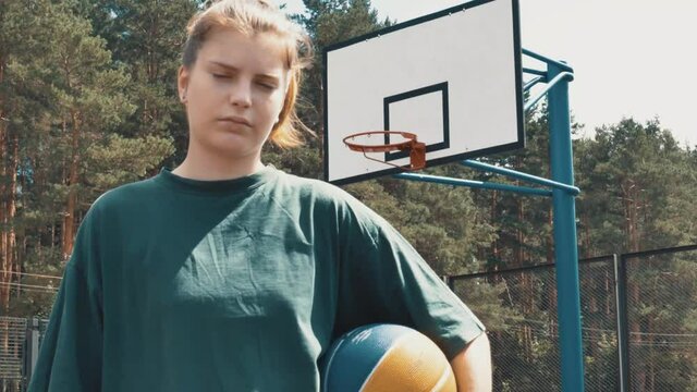 Portrait of attractive serious teenage female holding basketball ball looking at camera. Concept of sport, power, competition, active lifestyle. Girl player playing basketball on the court