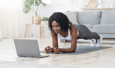 Fototapeta premium Black girl planking at living room, looking at laptop screen