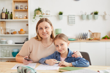 Warm toned portrait of happy mother and daughter smiling at camera while sitting at desk together...