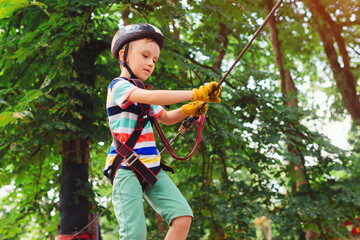 Naklejka na ściany i meble Summer camp. Boy passing the cable route high among trees. Climbing the rope park.