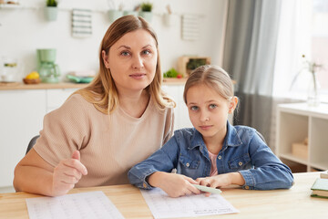 Warm-toned portrait of adult mother helping daughter doing homework while studying at home and looking at camera