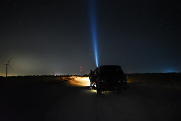 night photo. Dark silhouette of a man near a car on a dirt road with a flashlight shining in the...