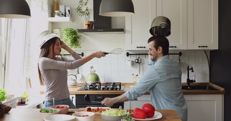 In modern home kitchen cheerful millennial couple take break from food preparation wear cookware holding kitchenware supplies fighting having fun laughing. Funny routine, happy family, cookery concept - Powered by Adobe