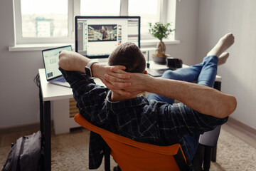 Man relaxing while working remotely from home holding legs on the table looking at the computer stretching hands behind his head