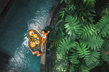 Travel happy couple in love eating floating breakfast in jungle swimming pool. Awakening in morning. Black rattan tray in heart shape, Valentines day or honeymoon surprise, view from above. - 366041533
