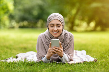 Pretty girl in hijab laying on grass, using phone